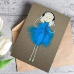 Handmade card for the phenomenal woman. A girl adorned with blue feathers is listing music. This card was made in the UK.
