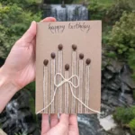 Bespoke and Personalised Handmade Card: Charm of Birthday Candles. Made of Coffee beans, cotton string on brown paper in front of waterfall.