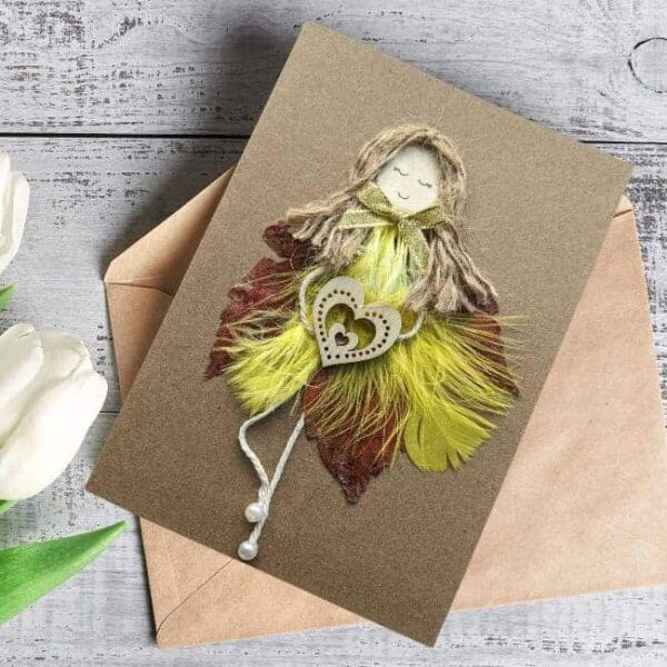 Best handmade romantic card for her shows a woman made of yellow feathers and leaves holding a wooden heart.