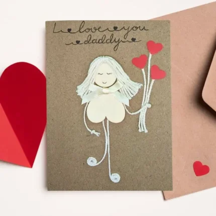 The Handmade and Heartfelt I Love You Dad Card features a girl made of wood with heart-shaped balloons on brown paper, complemented by red heart cut-outs for extra love.