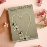A handmade Mother's Day card with "Thank You Mum" elegantly scripted alongside a heart and flowers crafted from rope and buttons, resting on a pink envelope. Delicate pink flowers are scattered around this heartfelt creation.
