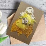 Beautiful Handmade Happy Birthday Card featuring a whimsical figure made of yellow and gold feathers and paper, holding a heart, set on a textured brown envelope.