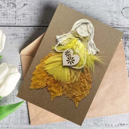 Beautiful Handmade Happy Birthday Card featuring a whimsical figure made of yellow and gold feathers and paper, holding a heart, set on a textured brown envelope.