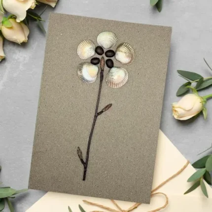 Nature-Inspired Birthday Card featuring a handmade flower design with seashell petals and twig stem, surrounded by white roses and green leaves.