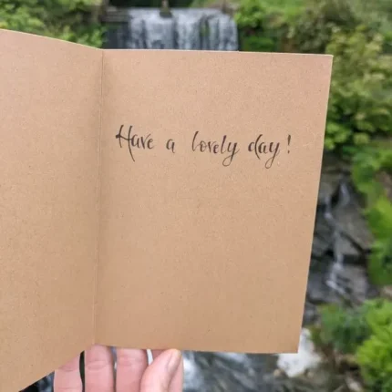 Handmade greeting card with elegant black calligraphy on kraft paper, photographed outdoors with a blurred natural background.