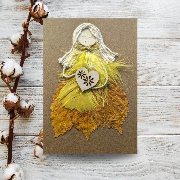 The Handmade Cards For Her category, image displays a card on a wood table featuring a figure with a yarn head, yellow feather body, and wooden heart, complemented by a cotton branch.