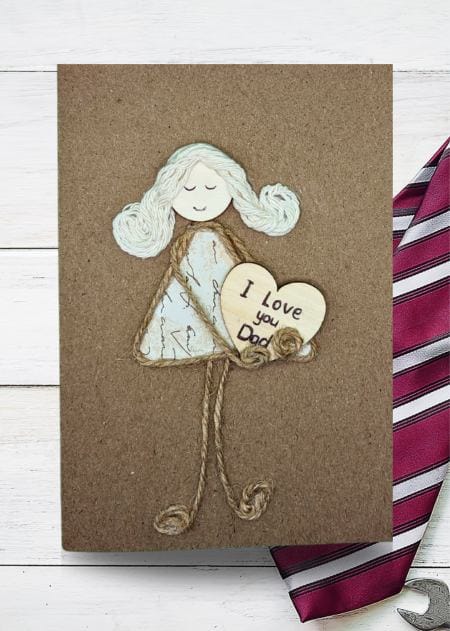 The handmade Father’s Day card features a girl holding a heart and a tool, symbolizing love and craftsmanship, and is part of a handcrafted collection.