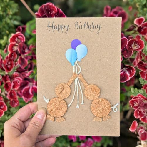 Cork in handmade card featuring a birthday design with cork figures holding balloons, photographed outdoors among red flowers.