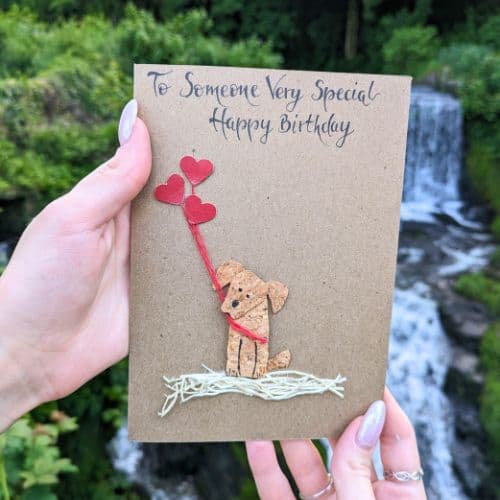 Cork in handmade card showing a teddy bear made from cork holding red heart balloons, photographed outdoors near a waterfall.