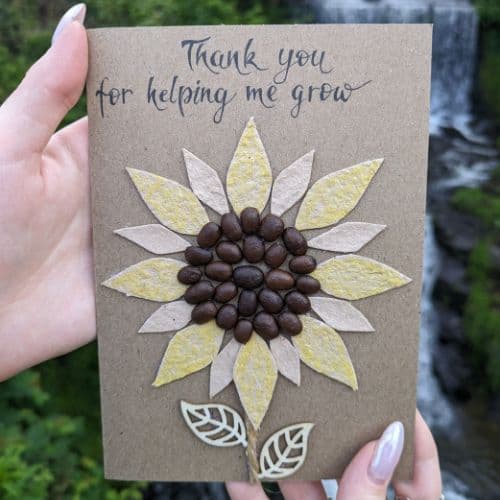 Coffee in handmade cards shown as a handmade thank you card with a sunflower design made from real coffee beans and layered paper petals, held outdoors near flowing water.