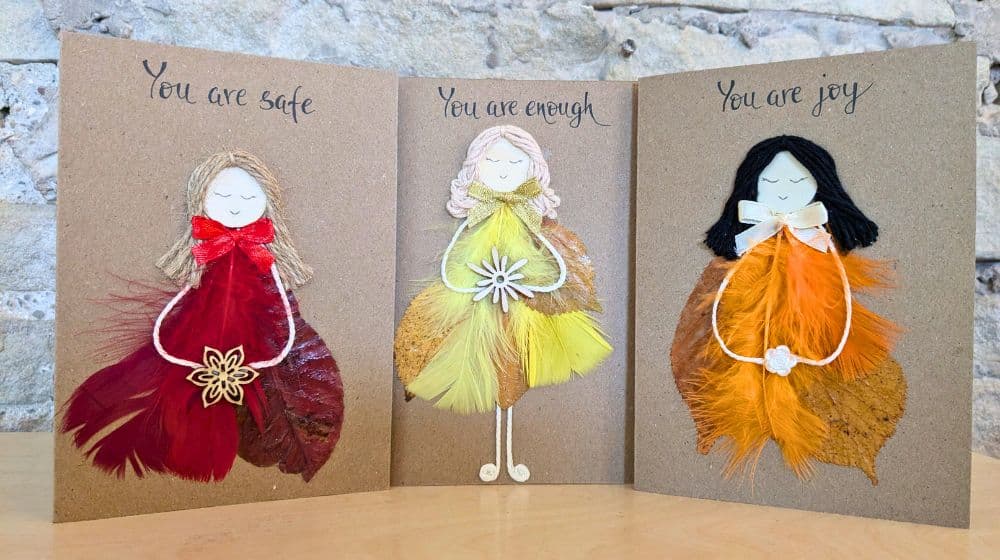 Feathers in handmade cards displayed as three handcrafted greeting cards with angel figures made from natural feathers and handwritten affirmations on kraft card.