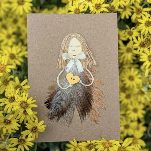 Feathers in handmade cards shown as a single handcrafted greeting card with an angel figure made from natural feathers, photographed among yellow flowers.