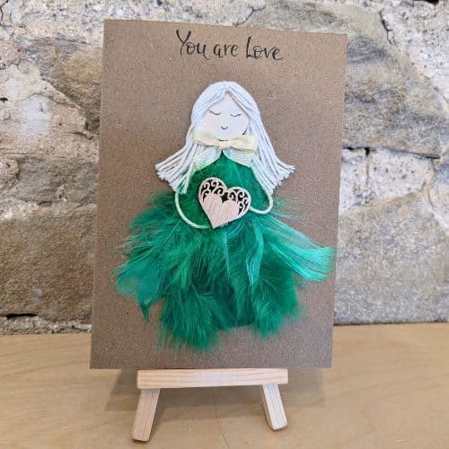 Feathers in handmade cards shown as a handcrafted greeting card with a green feather dress angel figure holding a heart, displayed on a small wooden stand.