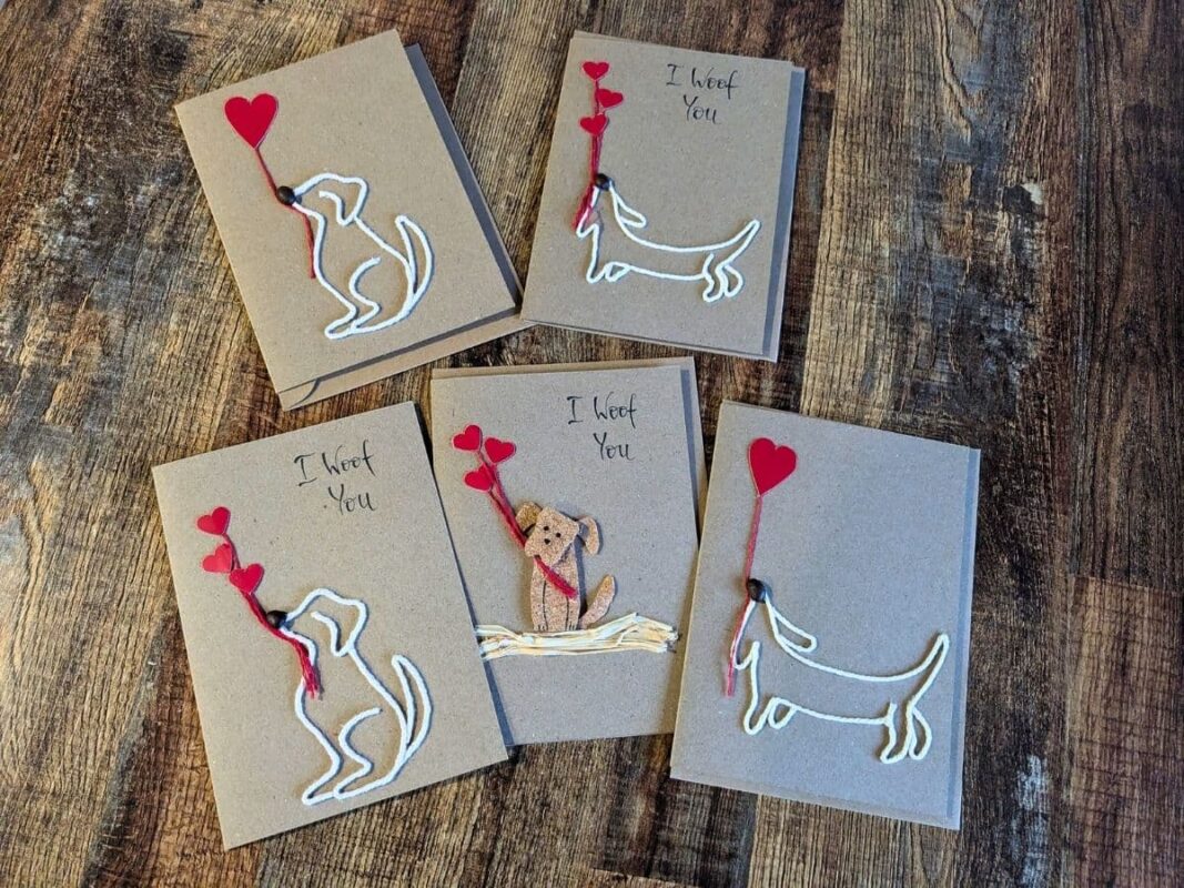 A collection of five handmade kraft cards on a dark wood background featuring dogs crafted from white yarn and cork, holding red heart balloons, showcasing the Gifting Psychology of artisan stationery.