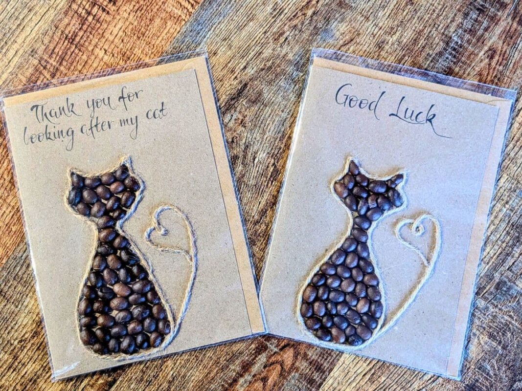 Two handmade better than store-bought cards featuring a sitting cat silhouette filled with real coffee beans and a twine heart tail, with handwritten messages for "Thank you for looking after my cat" and "Good Luck" on a wooden surface.