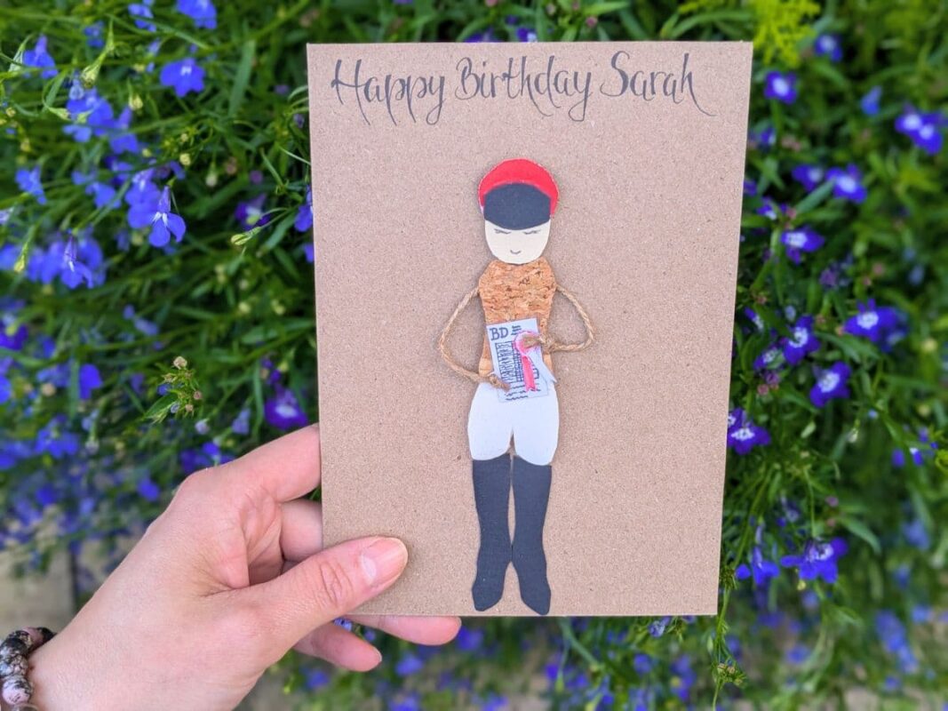 A hand holding a personalized equestrian-themed birthday card for Sarah, featuring a 3D rider figure with cork clothing and twine arms, illustrating why these are handmade better than store-bought cards.