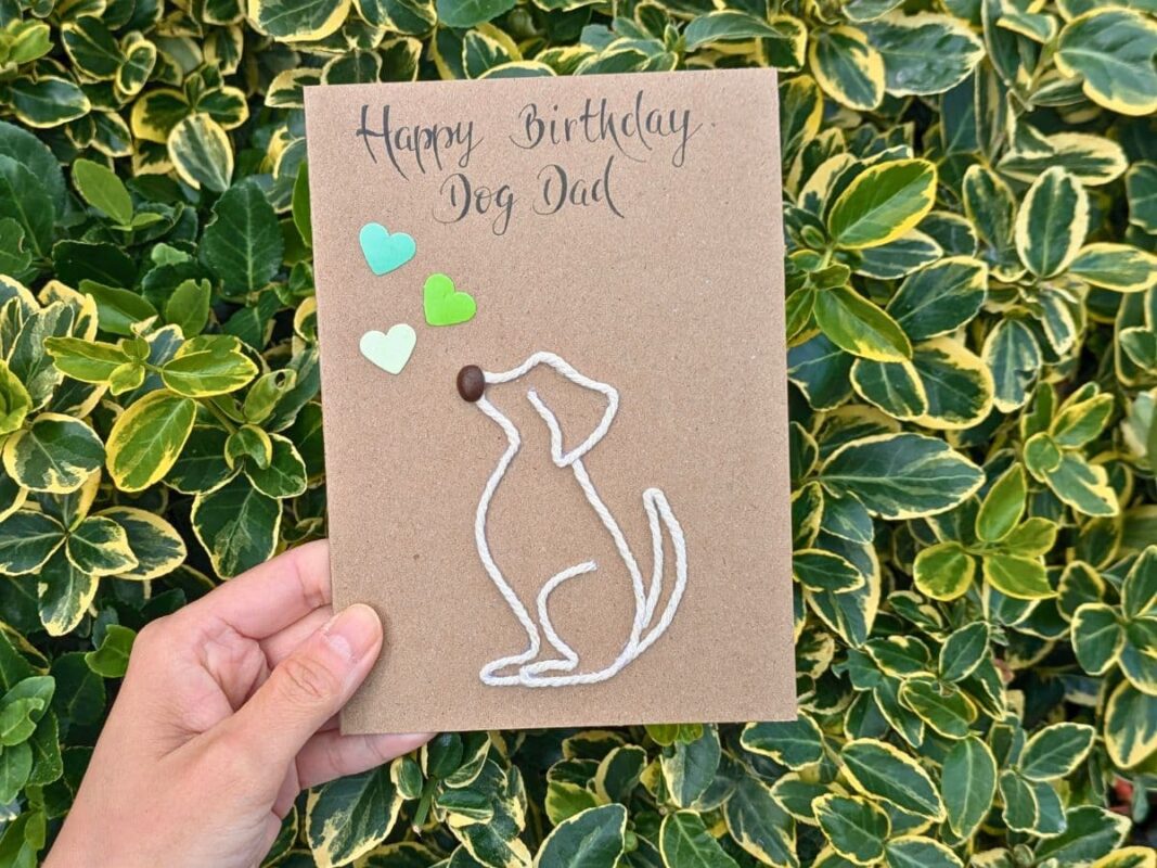 A hand holding a "Happy Birthday Dog Dad" card, featuring a minimalist dog outline made of white string, a coffee bean nose, and green paper hearts, demonstrating why these are handmade better than store-bought cards.