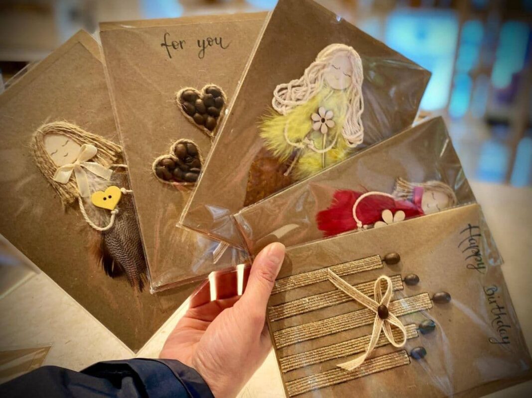 A hand holding a fan of five individually wrapped handmade better than store-bought cards, showcasing diverse designs including 3D figures with feathers and twine, coffee bean hearts, and a "Happy Birthday" card.