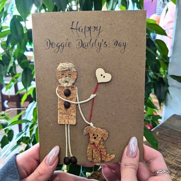 A "Happy Doggie Daddy's Day" kraft card featuring a man and his dog crafted from luxury handmade card materials like natural tree bark, cork, and twine, accented with a wooden heart button.