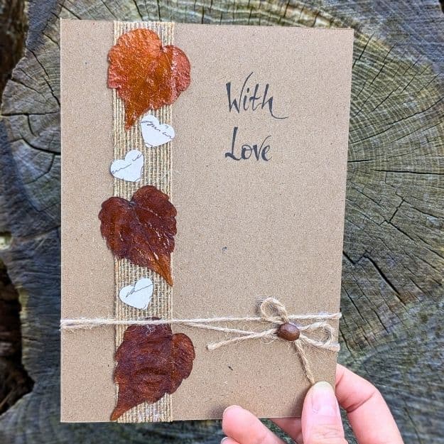A "With Love" kraft card featuring three natural dried leaves with a shimmering copper finish, accented by small white hearts and a twine bow, showcasing creative luxury handmade card materials.