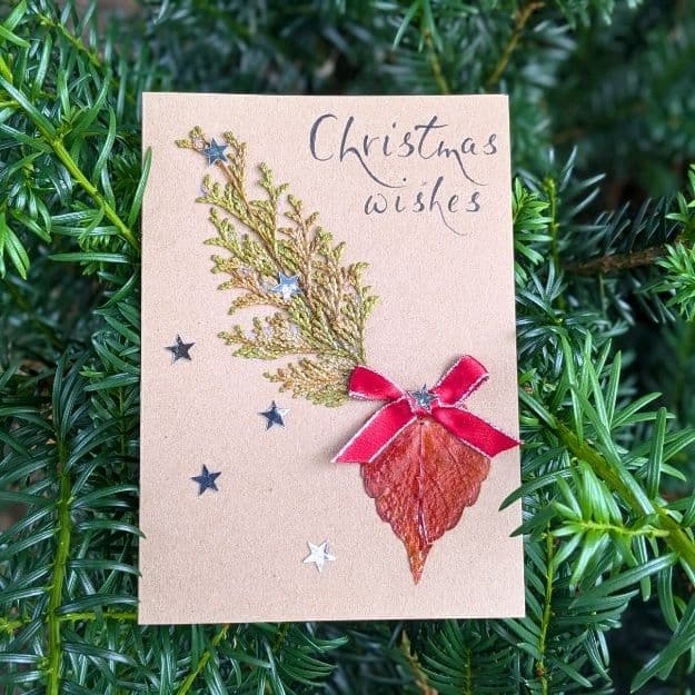 A "Christmas Wishes" kraft card featuring a festive tree design made from a cedar garland branch and a shimmering copper dry leaf, accented with silver stars and a red bow using luxury handmade card materials.