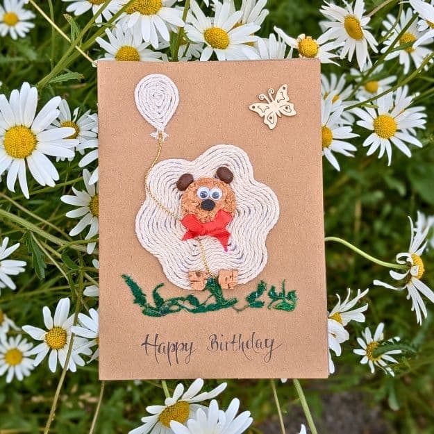 A "Happy Birthday" kraft card featuring a 3D poodle design crafted from white yarn, cork, and a red bow, showcasing the creative use of luxury handmade card materials against a backdrop of white daisies.