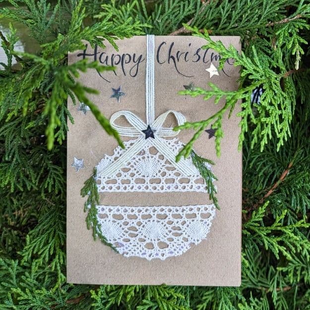 A "Happy Christmas" kraft card featuring a festive bauble made from delicate white lace, framed by green cedar sprigs and silver stars, created with luxury handmade card materials.