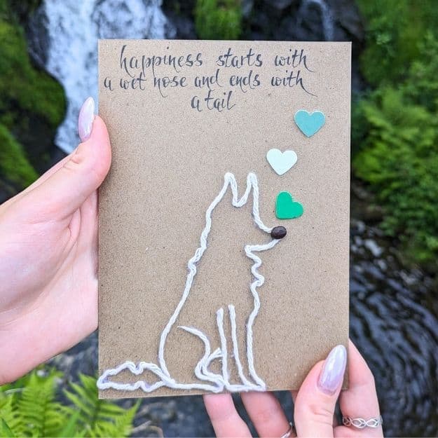A portrait-oriented kraft card featuring handmade calligraphy that reads "happiness starts with a wet nose and ends with a tail," featuring a white yarn dog silhouette and green heart accents.