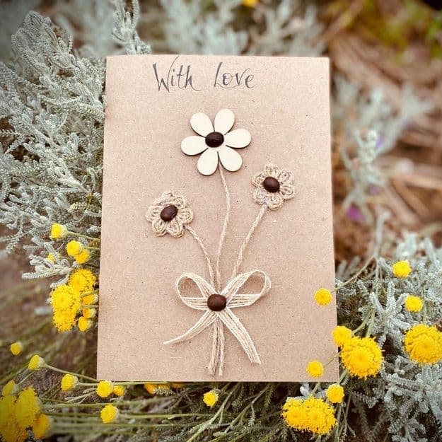 Handmade card with floral motifs crafted from luxury handmade card materials, including wooden cutouts, jute twine, and coffee beans, displayed among yellow wildflowers.