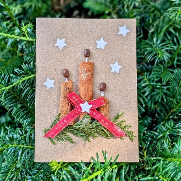 A festive Christmas card featuring candles made from cinnamon sticks and coffee bean flames, embellished with a red ribbon and white stars using luxury handmade card materials.