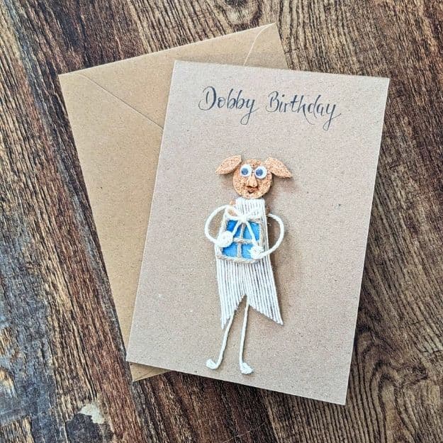 A whimsical kraft birthday card featuring a character crafted from luxury handmade card materials like textured cork and white cotton twine, shown with a matching 5 x 7" recycled envelope on a wooden surface.