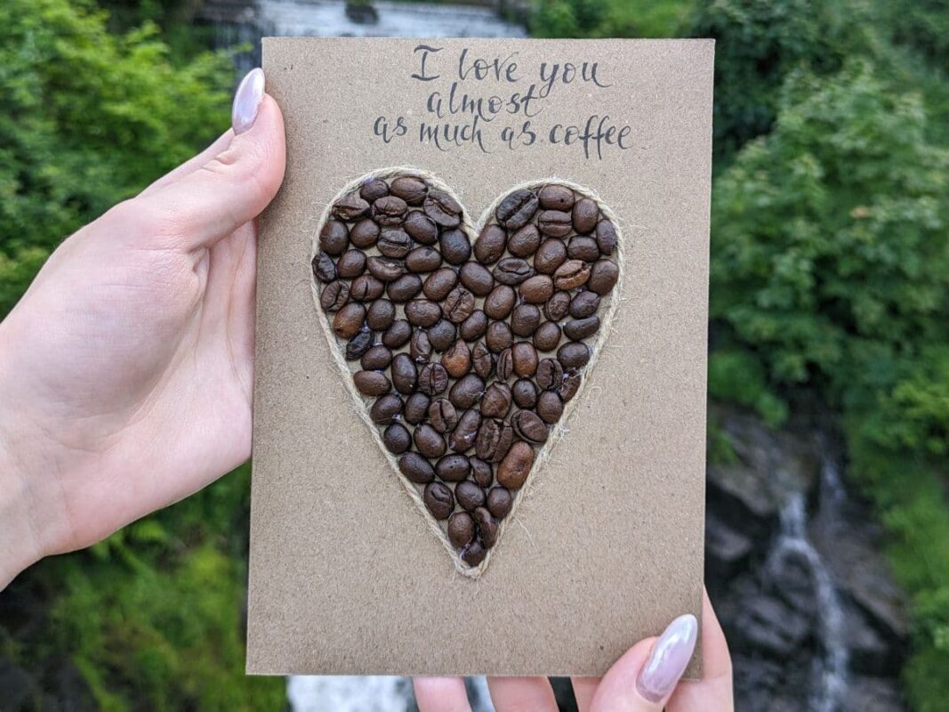 A romantic handmade Valentine’s card UK from featuring a heart shape made of real coffee beans outlined with twine on brown kraft card, with handwritten text saying "I love you almost as much as coffee.