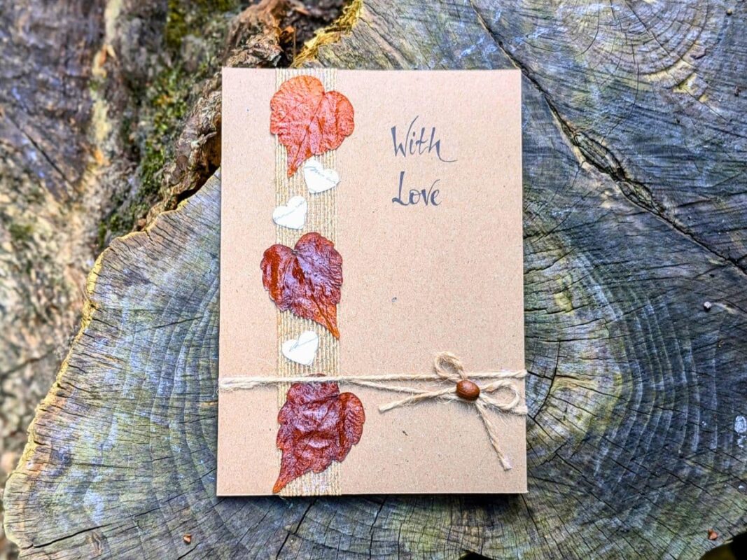 A romantic handmade Valentine’s card UK displayed on a tree stump, featuring three pressed autumn leaves and small white heart cards on a burlap strip, finished with a twine bow and the handwritten message "With Love."