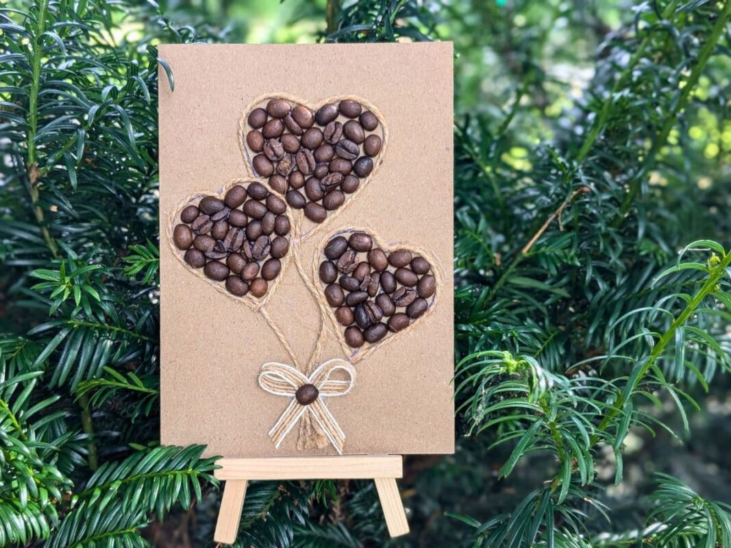 A romantic handmade Valentine’s card UK for coffee lovers featuring three heart cards shaped like balloons made from real coffee beans, outlined with twine and finished with a rustic bow, displayed on a wooden easel against a green garden background.