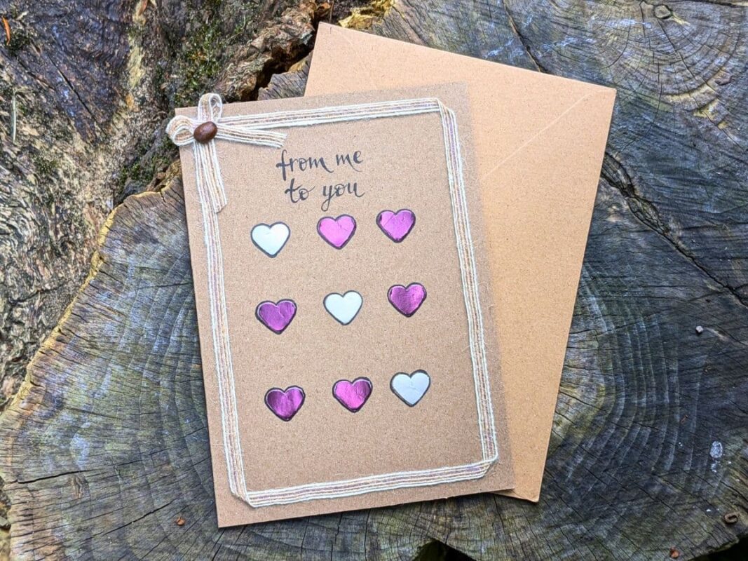 A romantic handmade Valentine’s card UK featuring a grid of nine pink and white heart cards on kraft paper with a twine border, a coffee bean bow detail, and handwritten text reading "from me to you," displayed on a rustic wood background with a matching envelope.